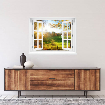 Erleben Sie die herbstliche Idylle in Ihren eigenen vier Wänden! Selbstklebendes Wandtattoo mit gedrucktem Bild von einem geöffneten Fenster mit Blick auf eine herbstliche Waldlichtung mit einer grünen Wiese und Sonnenstrahlen, die durch die Bäume scheinen, platziert über einem Sideboard in einem Wohnzimmer.